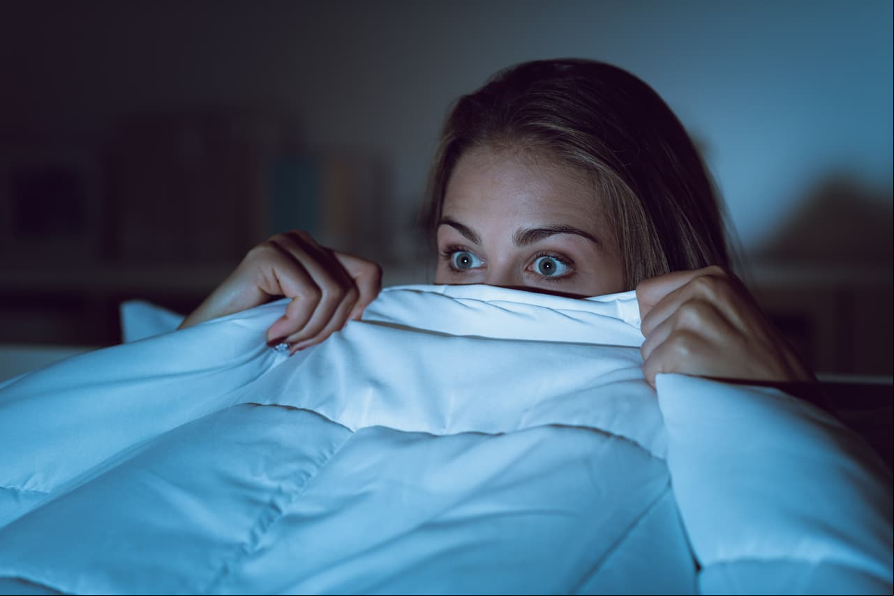 Woman watching a scary horror movie on tv late at night, she is frightened and hiding under the blanket