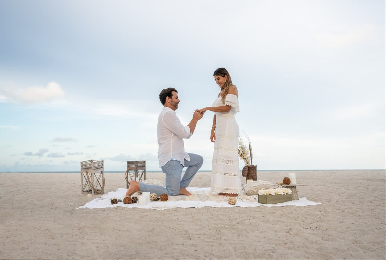 Loving couple getting engaged on a romantic picnic at the beach and him on his knees while he puts the ring on her finger - relationship concepts