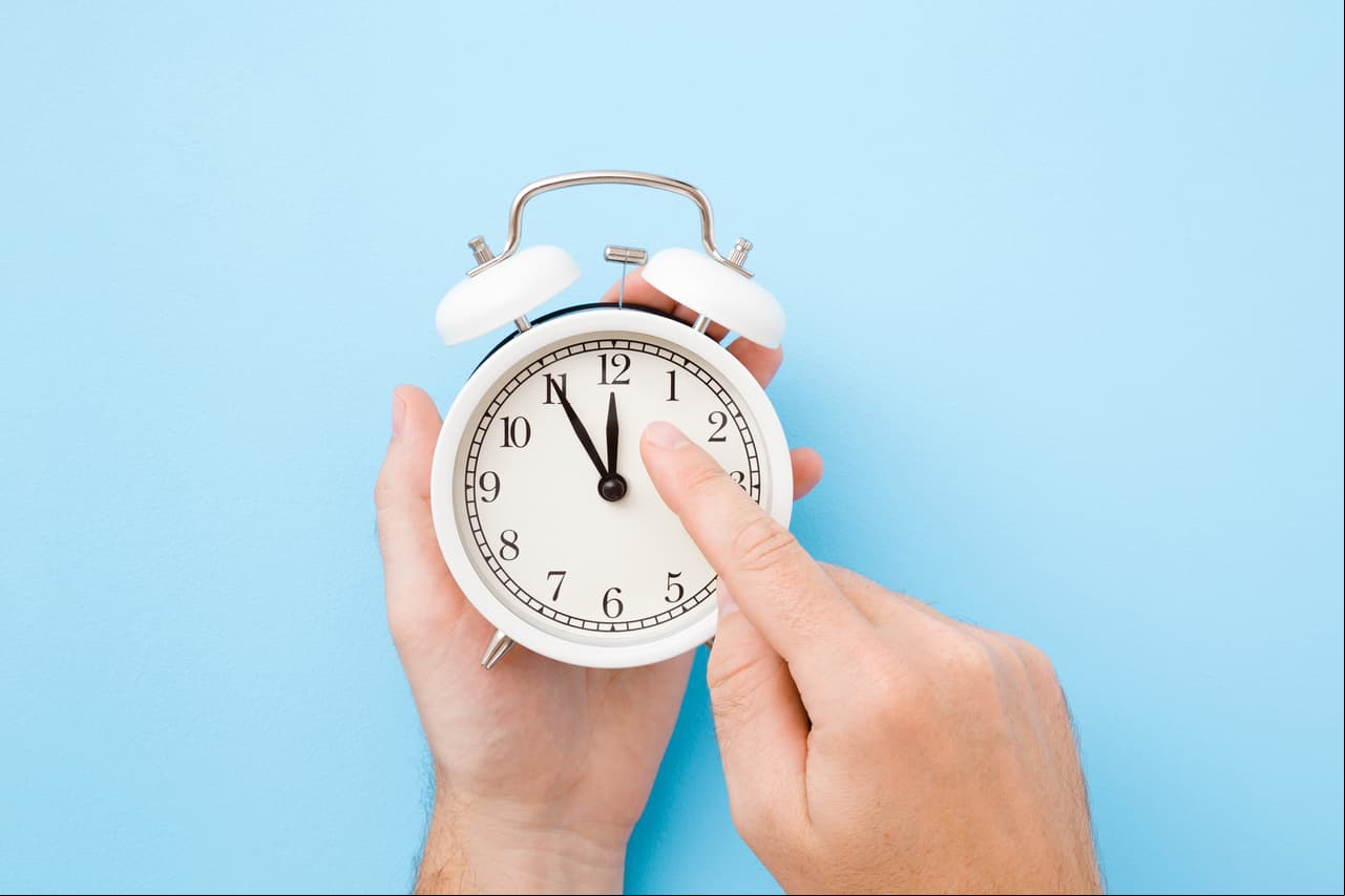Man hand holding white alarm clock. Finger pointing to arrow of twelve o'clock. Light pastel blue background. Time change concept. Closeup. Top view.