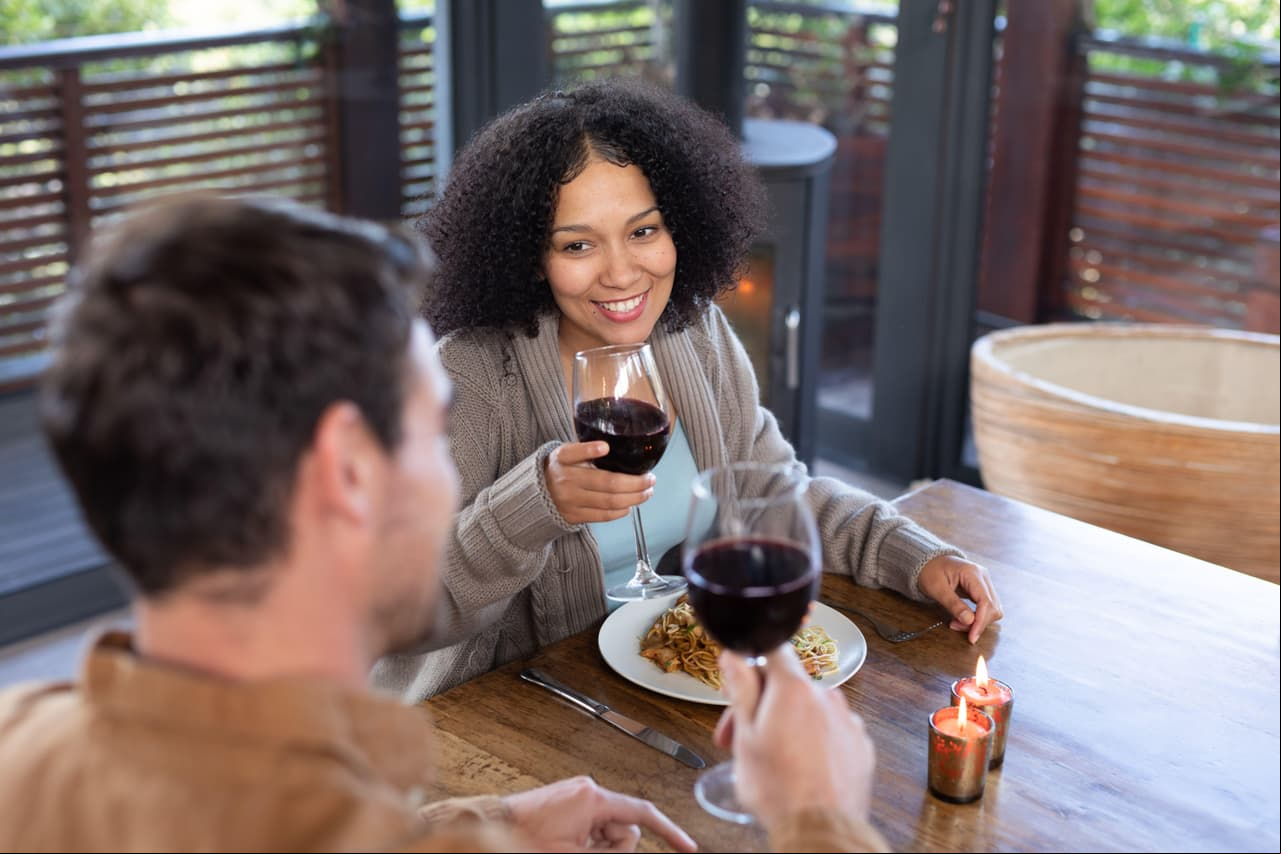 Happy diverse couple in living room sitting at table eating dinner together and drinking wine